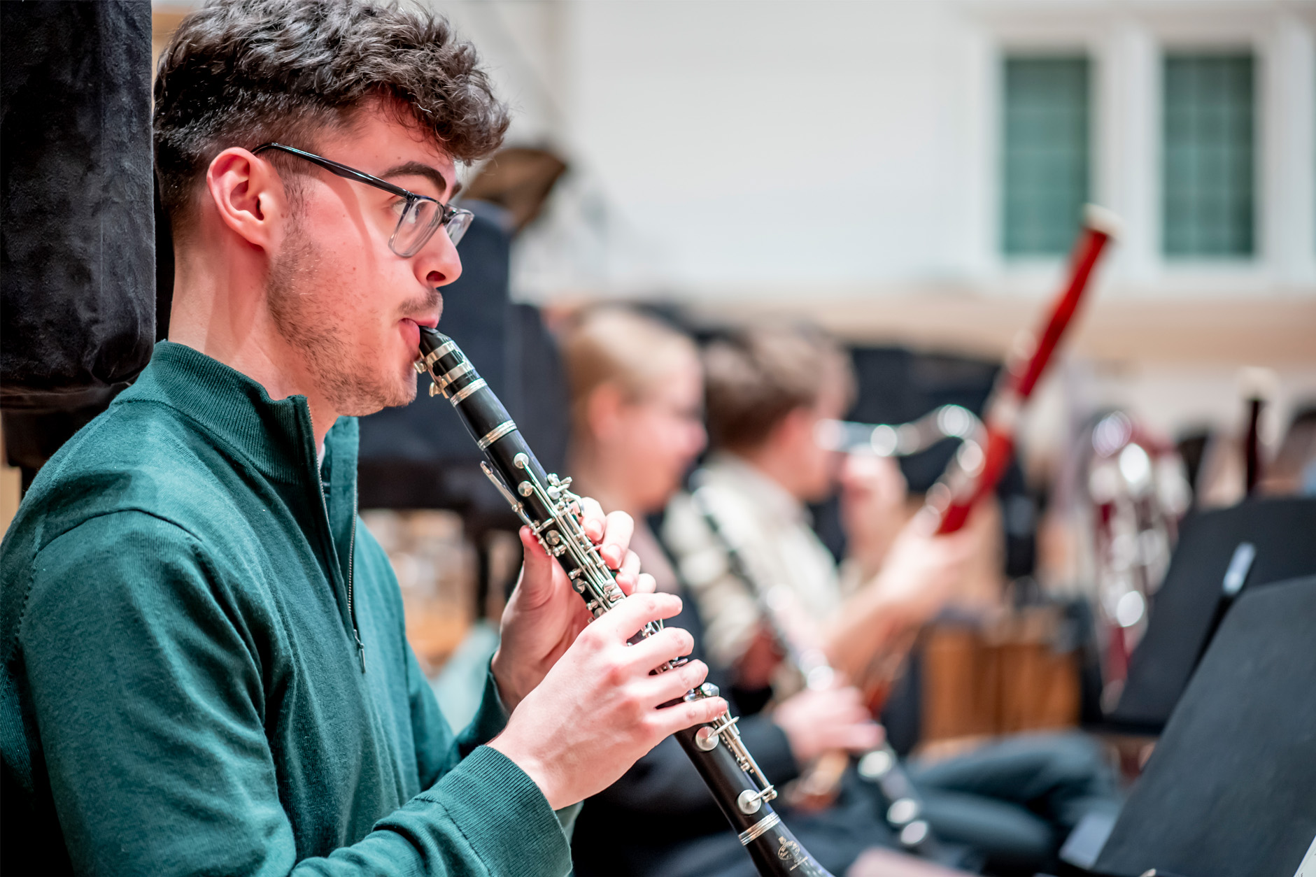 A male student wearing a teal jumper, playing the clarinet in an orchestral rehearsal.